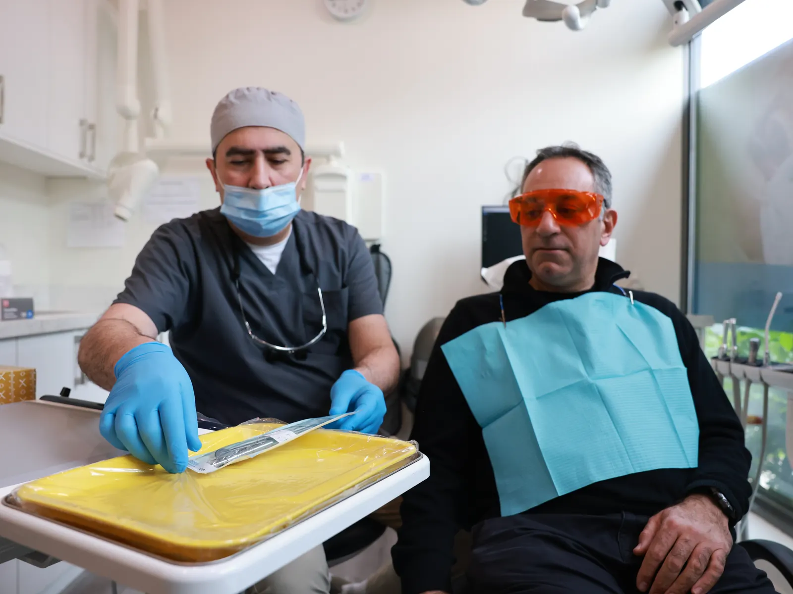 Dentist in scrubs and a mask explaining instruments to a seated patient wearing protective glasses before treatment